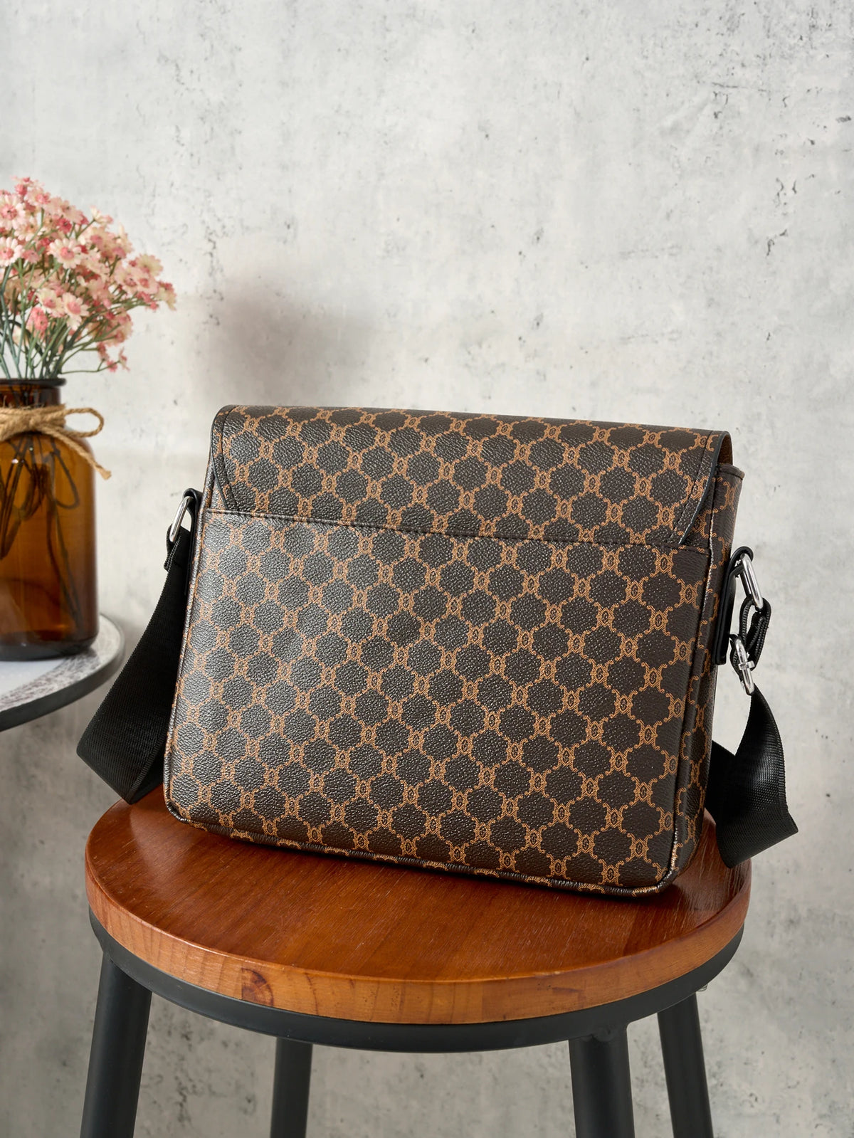 A brown patterned crossbody bag resting on a wooden stool next to a vase of pink flowers.