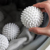 A hand holds a white reusable laundry ball with a textured surface, placed in a gray fabric container.