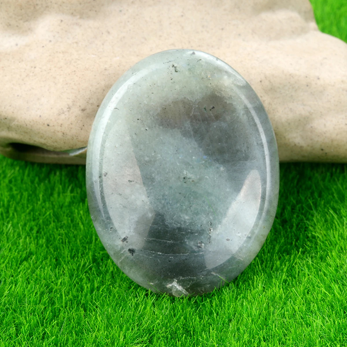 A smooth, oval gray natural crystal stone massage tool resting on bright green grass.