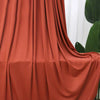 A close-up of flowing, rust-colored stretch jersey fabric draped in soft folds.