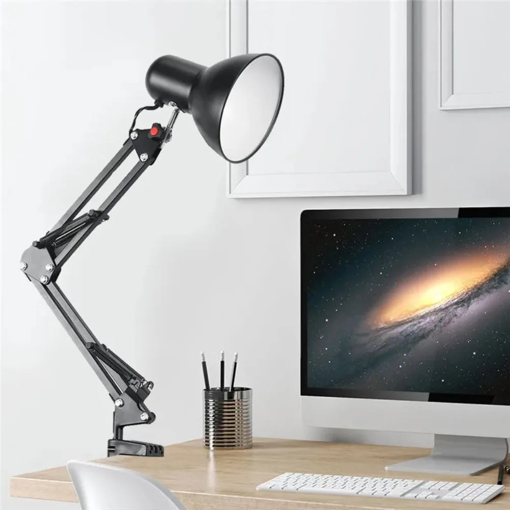 A black adjustable swing arm desk lamp is positioned next to a computer monitor on a wooden desk.