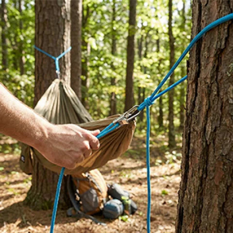 A hand is holding a burlap sack tied with blue rope, demonstrating a heavy-duty rope tensioner and tying tool in a forest set