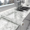 A marble peel-and-stick film is shown rolled out on a white marble countertop next to a kitchen sink.