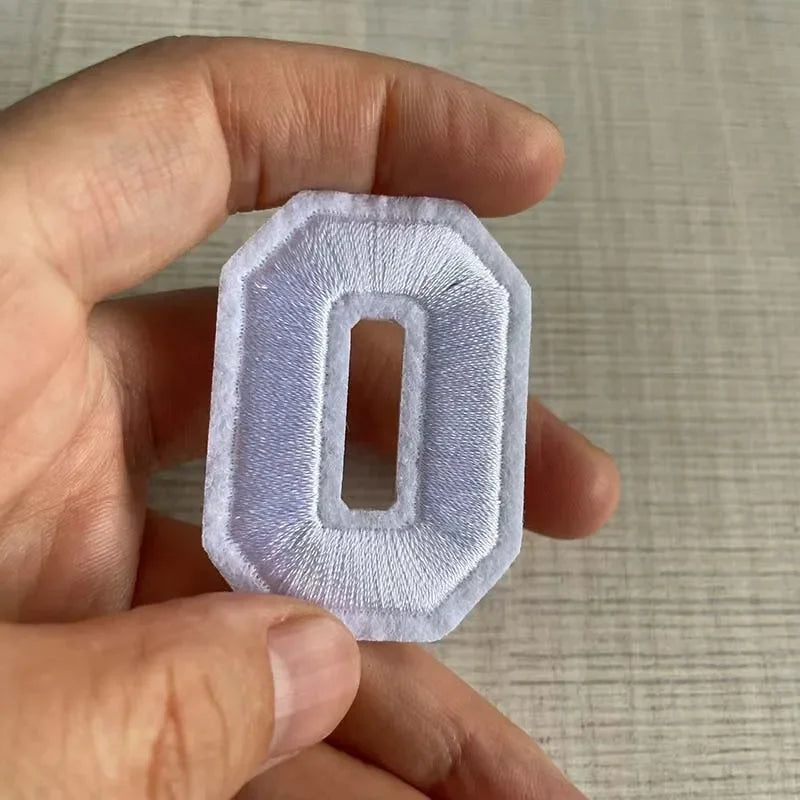 A hand holds a white iron-on 3D number patch shaped like the digit zero.