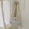 A white handwoven knit tote bag with a hollow design featuring blue floral embroidery, hanging on a white door frame.