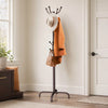 A freestanding metal coat rack with 14 hooks displaying a tan coat and a brown leather bag in a bright room.
