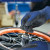 A hand wearing a blue work glove is using a tool to adjust a bicycle wheel.