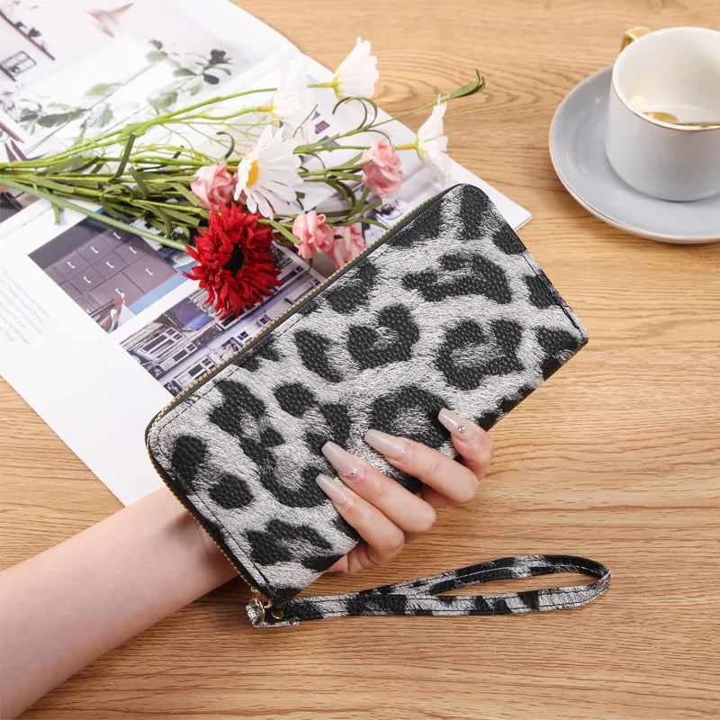 A hand holds a black and white leopard print retro long wallet on a wooden table next to some flowers and papers.