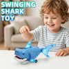 A smiling toddler is playing with a blue plastic swinging shark toy on a white table.