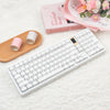 A white mechanical keyboard with pink keycaps is displayed next to a wooden tray and a pink book.