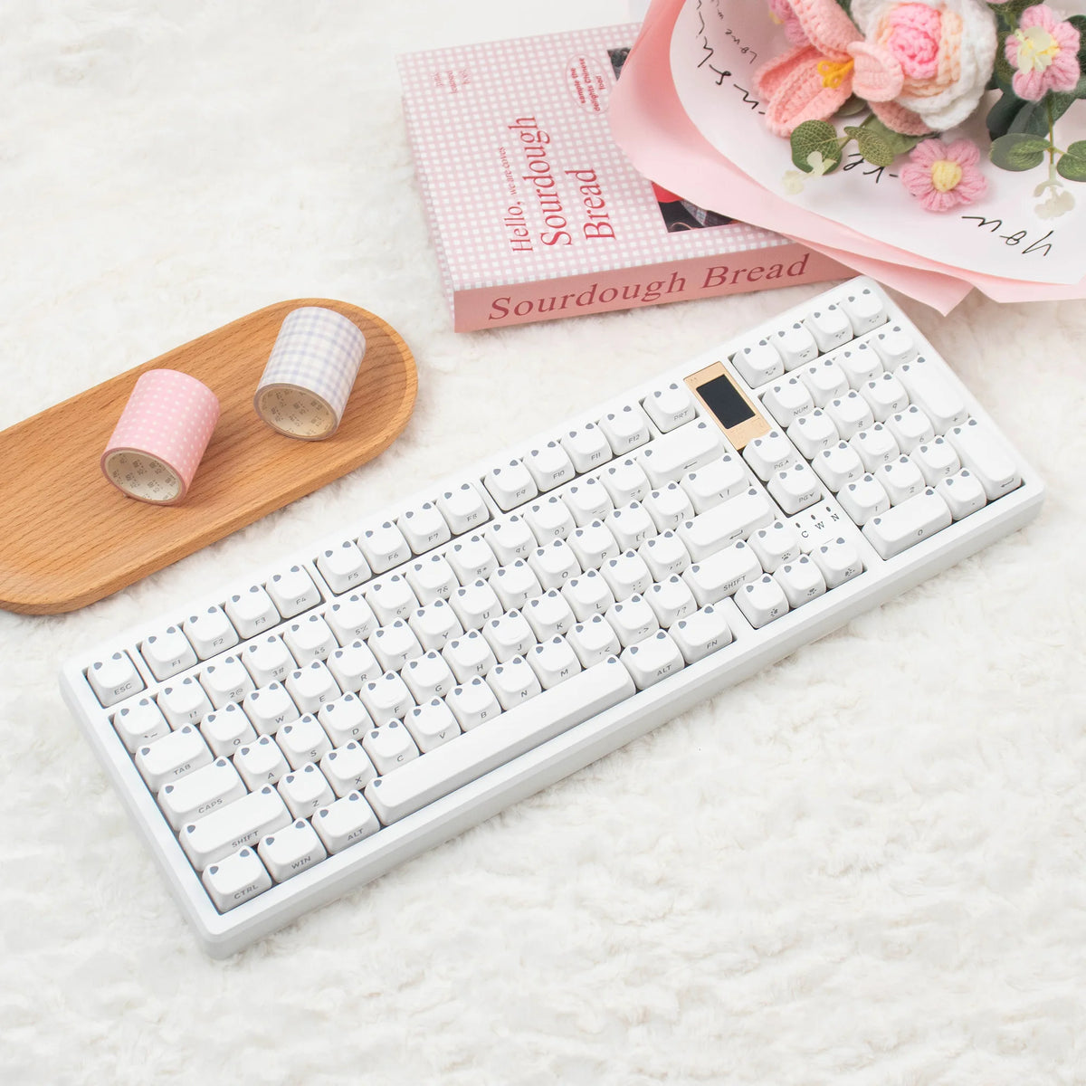 A white mechanical keyboard with pink keycaps is displayed next to a wooden tray and a pink book.
