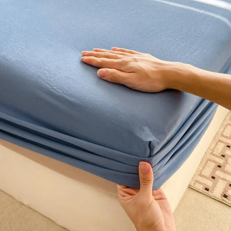 A hand is folding a blue cotton fitted sheet with an elastic band on a bed.