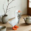 A white resin goose key holder sculpture with orange feet and a silver key hanging from its beak on a wooden desk.