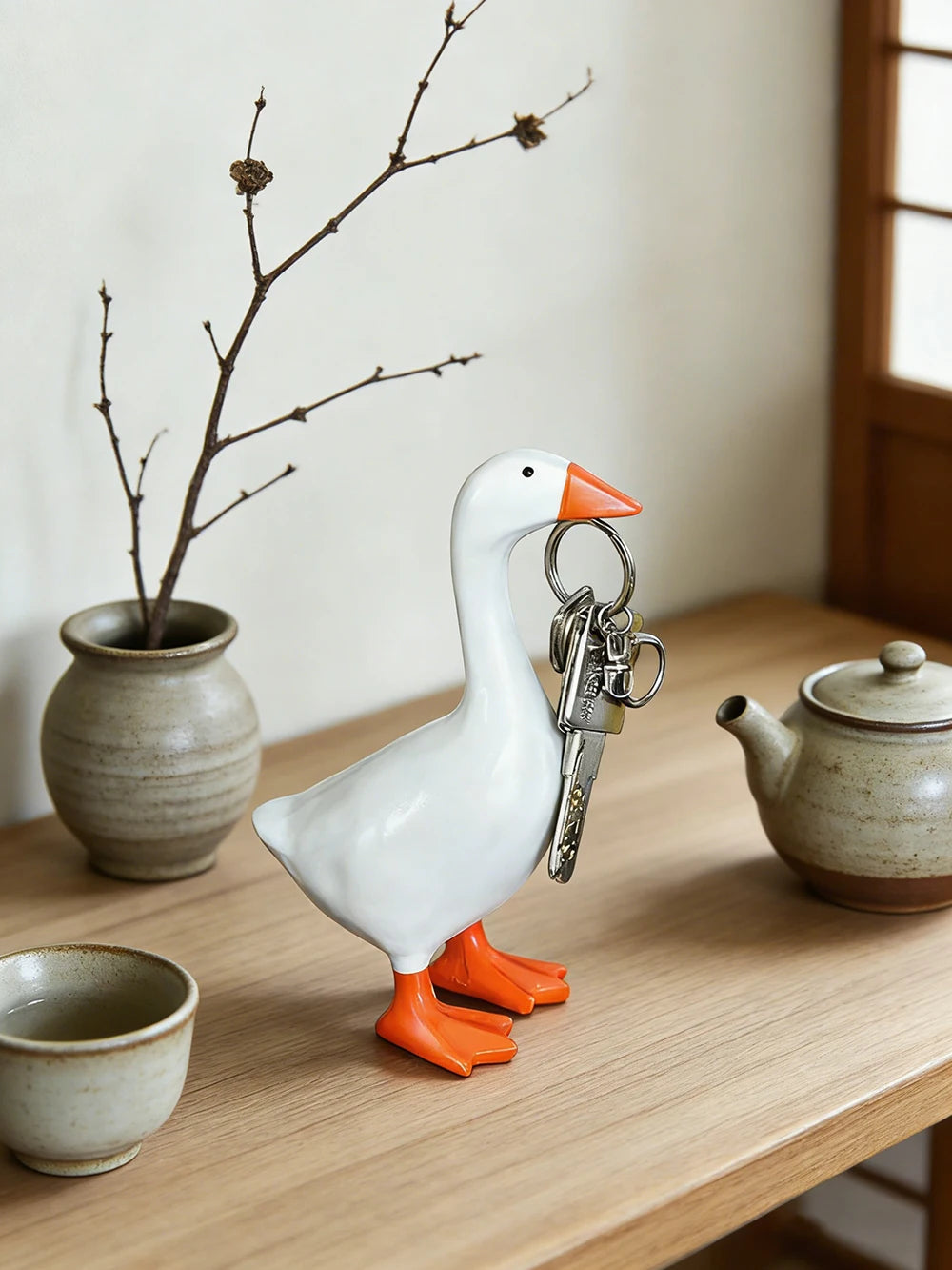 A white resin goose key holder sculpture with orange feet and a silver key hanging from its beak on a wooden desk.