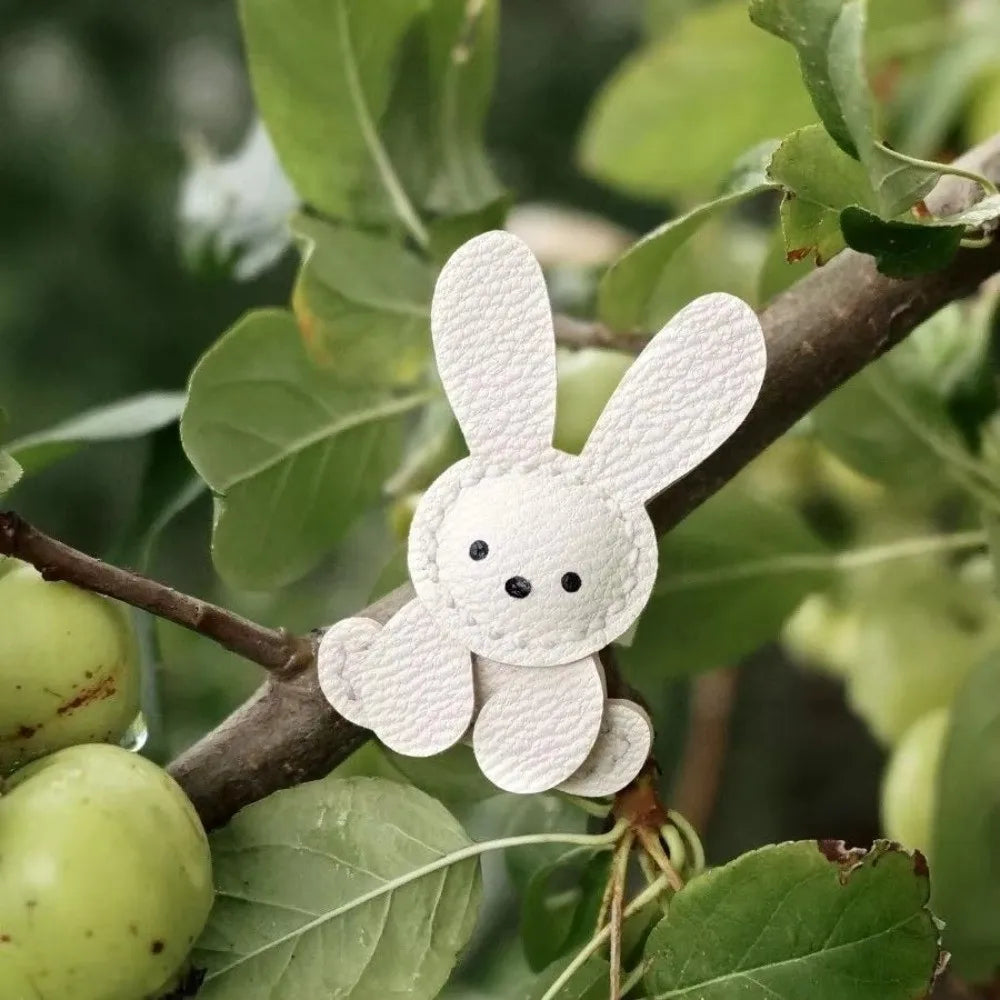 A white bunny bag charm keychain is attached to a tree branch surrounded by green leaves.