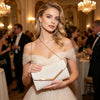 A woman wearing a beige pleated glitter envelope clutch with a gold chain strap at an elegant event.