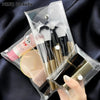 A transparent PVC cosmetic bag holds makeup brushes and a gold-colored cosmetic product on a dark background.