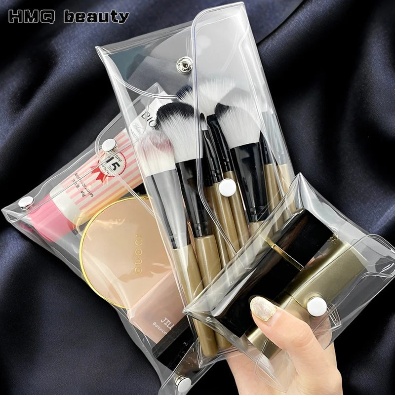 A transparent PVC cosmetic bag holds makeup brushes and a gold-colored cosmetic product on a dark background.