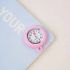 A pink silicone pocket watch with a brooch pin is placed on a light blue notebook.