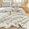 A light beige quilt with a gray floral pattern is displayed on a bed, featuring a white rabbit pillow and a green mug.