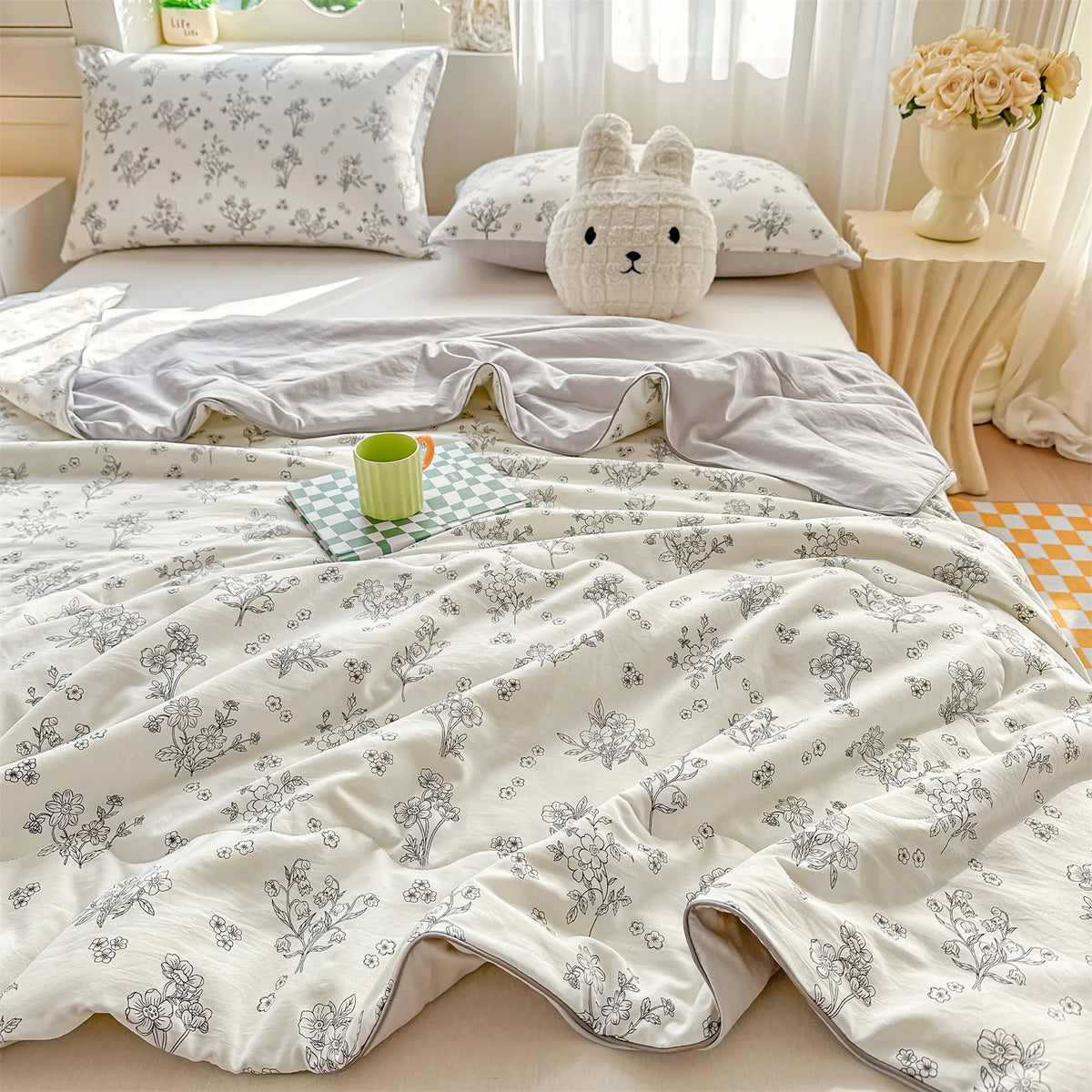 A light beige quilt with a gray floral pattern is displayed on a bed, featuring a white rabbit pillow and a green mug.