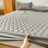 A gray waterproof fitted mattress protector is shown on a bed, with pillows and wooden furniture in the background.