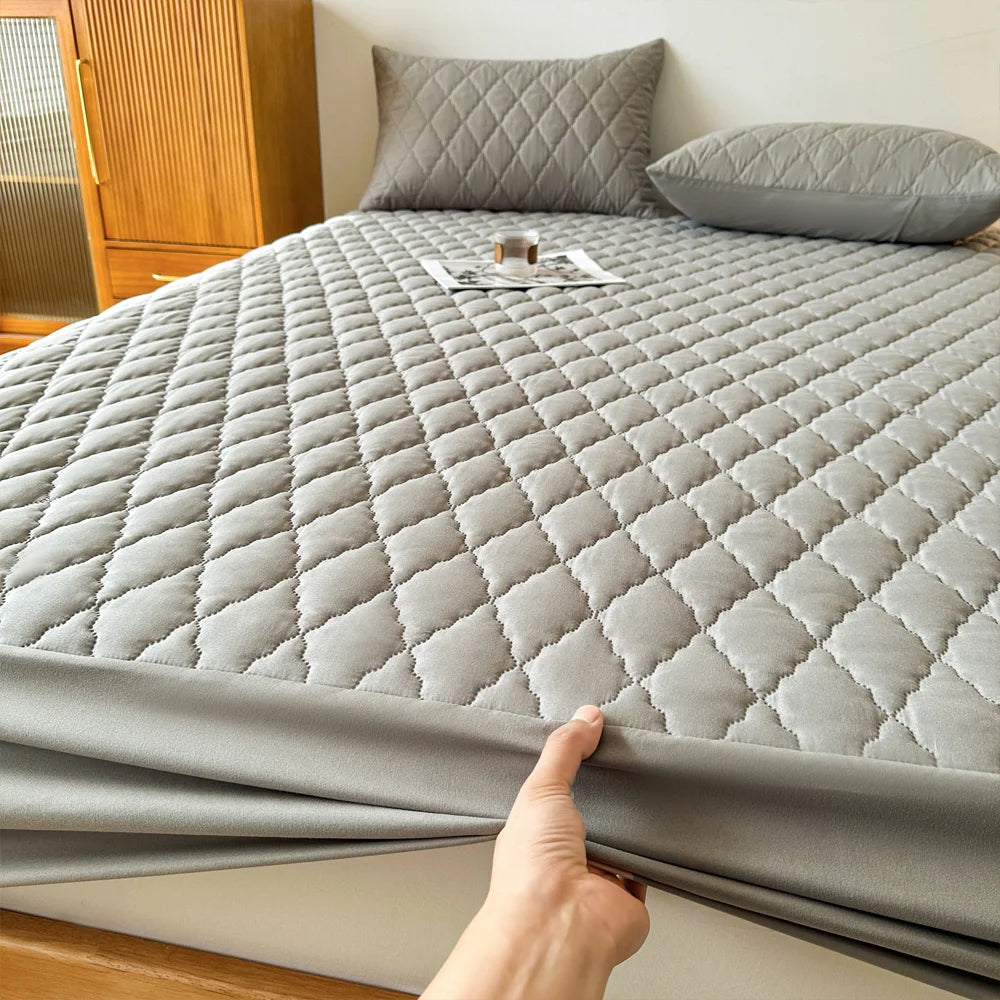 A gray waterproof fitted mattress protector is shown on a bed, with pillows and wooden furniture in the background.