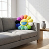 A cheerful sunflower plush cushion with a smiling face in various bright colors sits on a gray sofa.