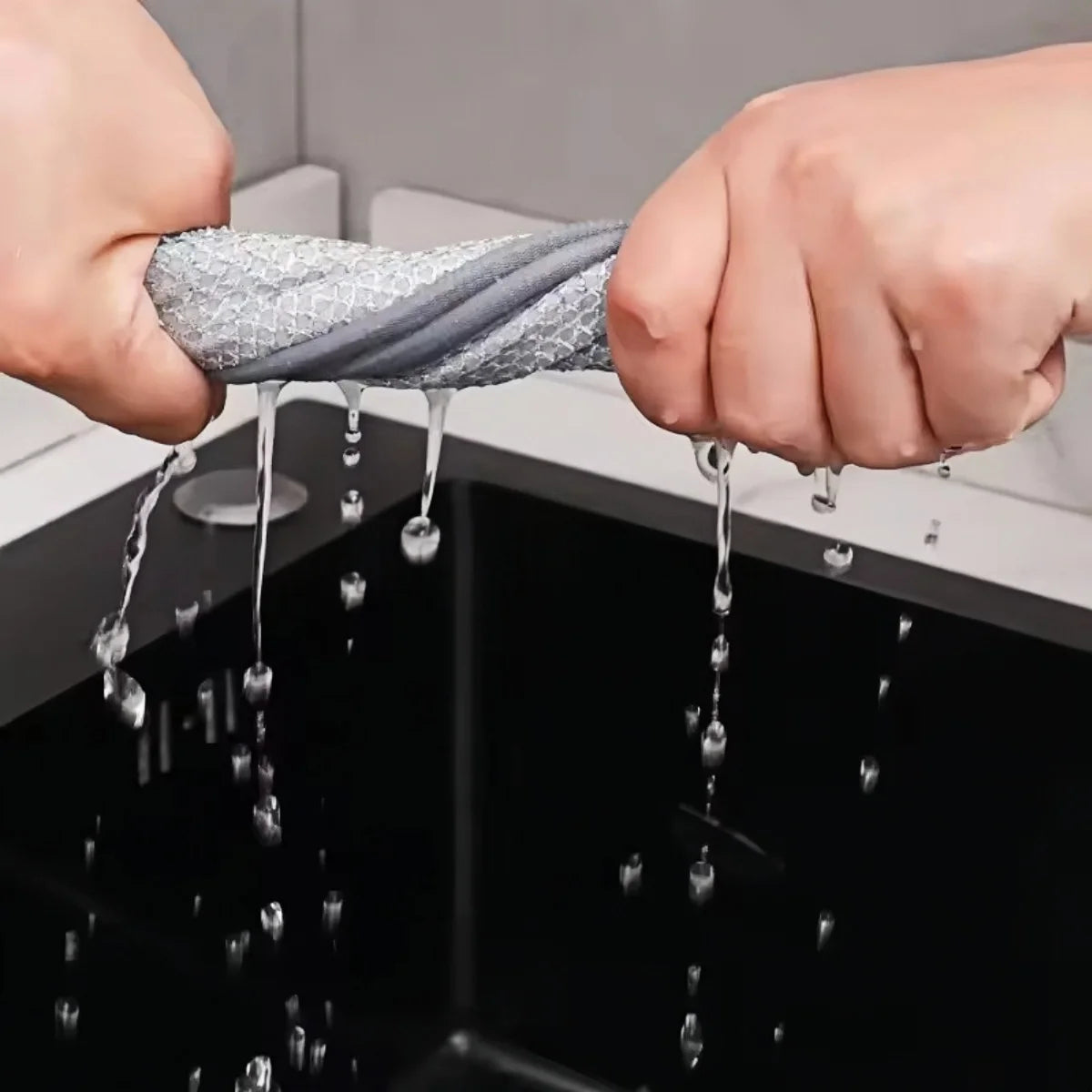 Hands are dripping water onto a black sink, showing a gray microfiber scouring cloth being used for dishwashing.
