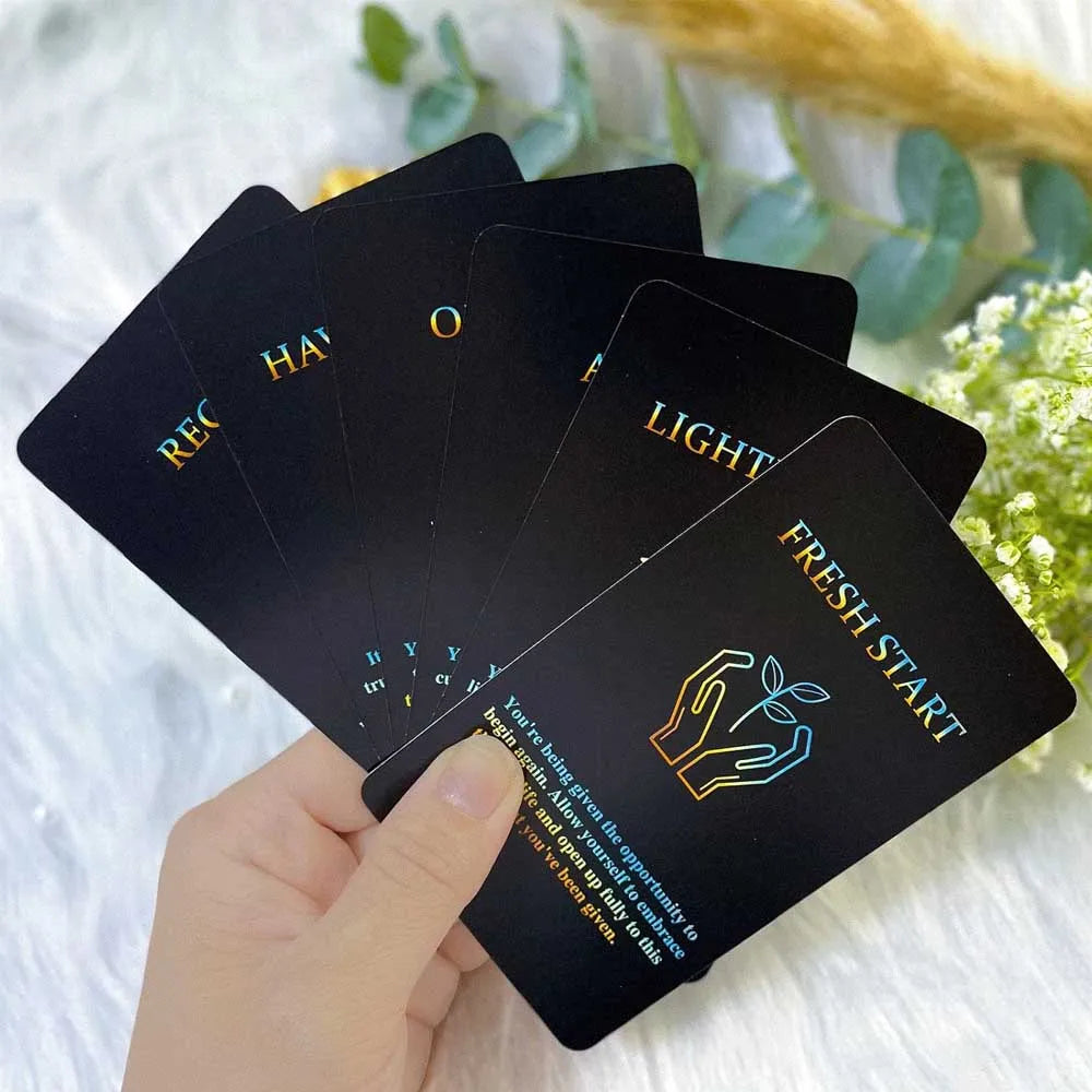 A hand holds a deck of oracle cards with titles like Recognition, Light, and Fresh Start against a blurred green background.
