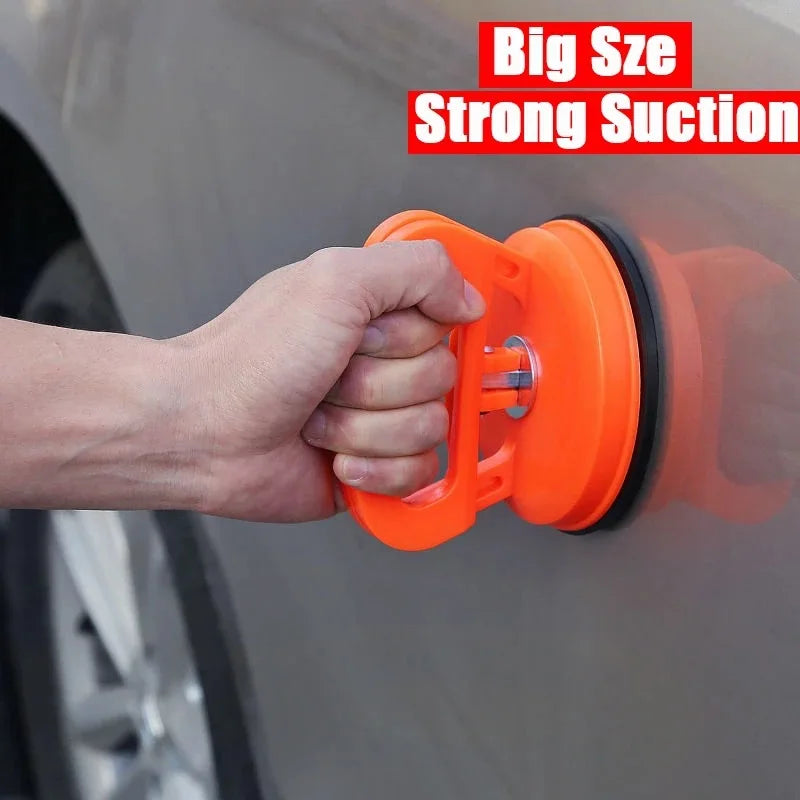 A hand is using an orange suction cup tool to pull a dent from a car body panel.