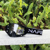 A pair of black HD Motocross goggles with an anti-fog lens resting on a light surface amidst green foliage.