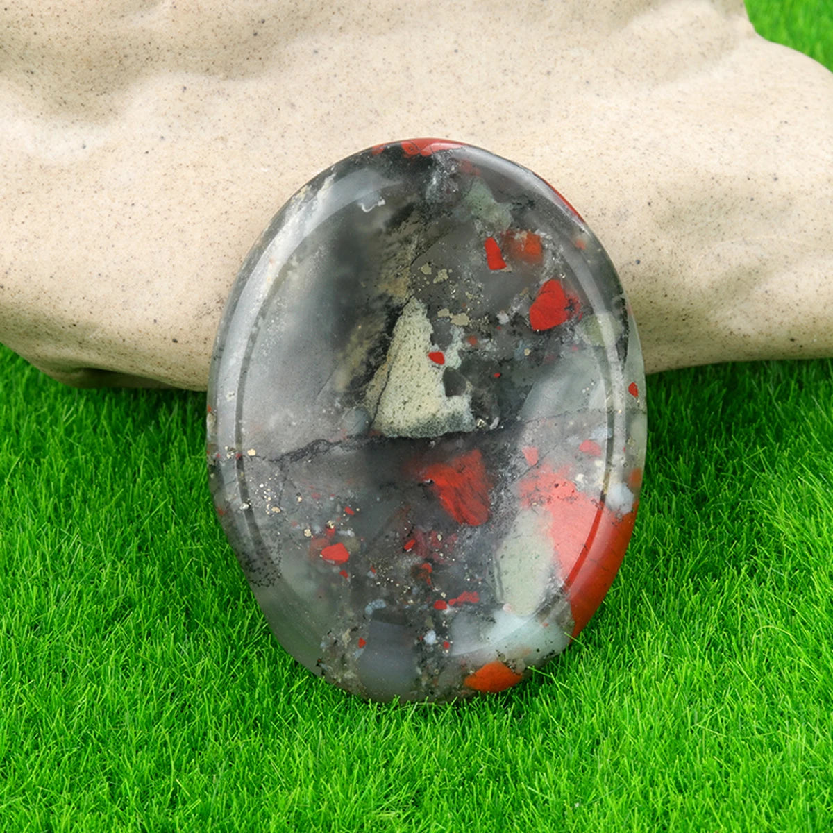 A polished, oval natural crystal stone with black and red inclusions rests on green grass.