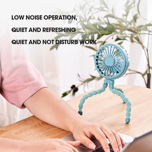 A hand is using a portable blue folding desktop fan on a wooden table next to a laptop.