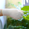 A hand wearing a disposable white nitrile glove gently touches a green leaf in a bright, indoor setting.