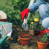 A person wearing red gloves is using a small shovel to plant seedlings in terracotta pots in a garden setting.
