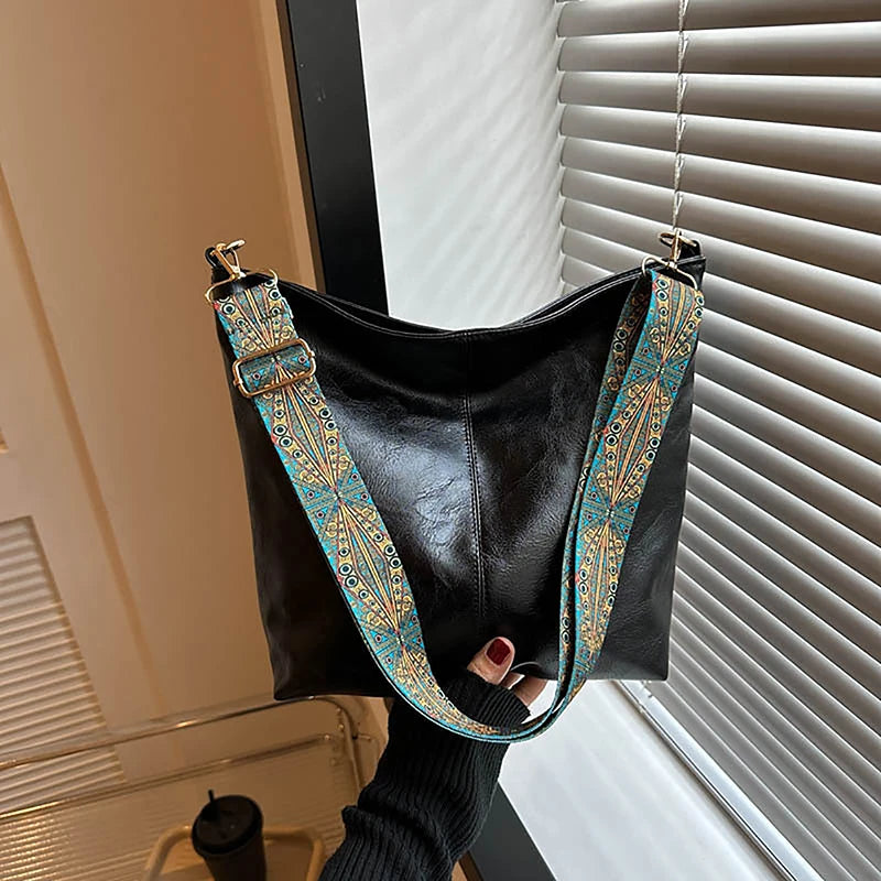 Sac à épaule noir à bretelles géométriques larges avec broderies or et sarcelle, accroché devant un store.