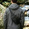 A person wearing a gray jacket is shown from the back, carrying a black sling bag on their shoulder in an outdoor setting.