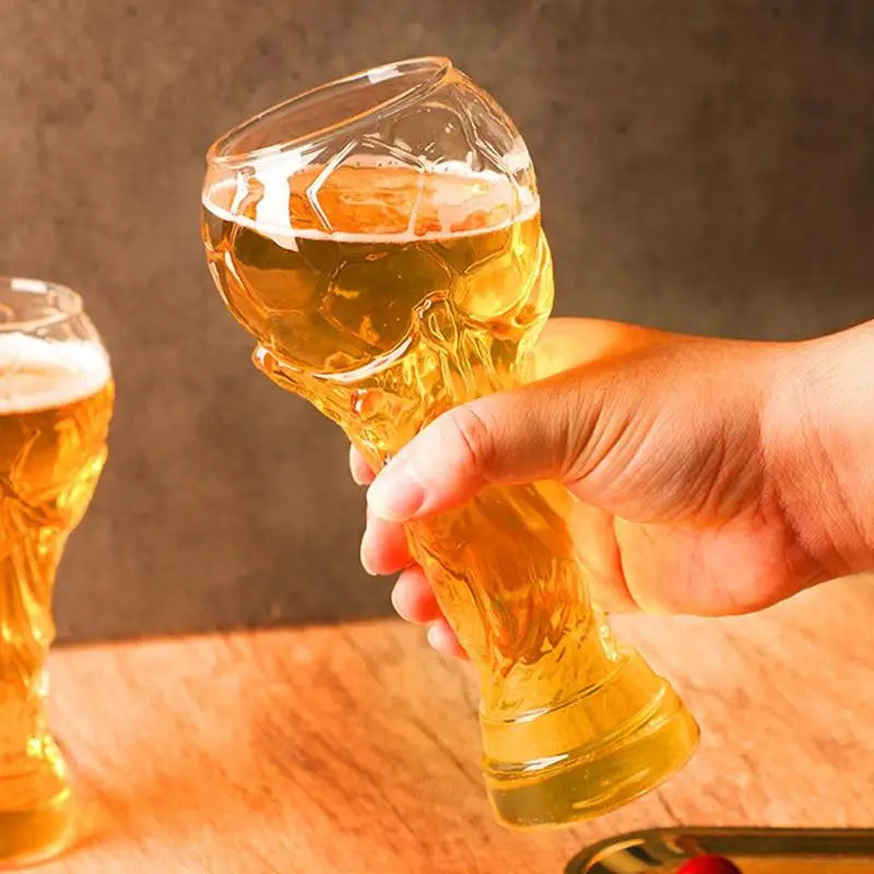 Eine Hand hält ein trofeenförmiges Glas, gefüllt mit goldenem Bier, auf einer Holzoberfläche.