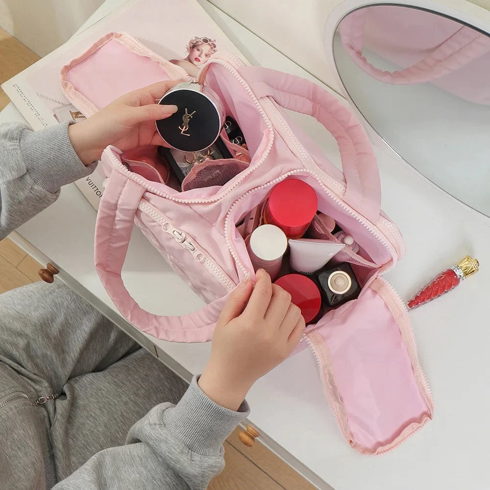 A hand is opening a pink travel makeup bag revealing various cosmetic products on a white surface.