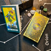A gold foil tarot card is displayed on a dark surface next to a guidebook and celestial map.