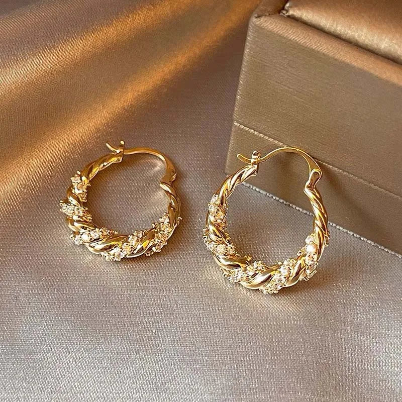 A pair of gold-finished twisted zircon hoop earrings resting on a beige fabric surface.