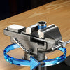 A close-up of a metal clamp-on vise with a blue rotating mechanism mounted on a wooden workbench.