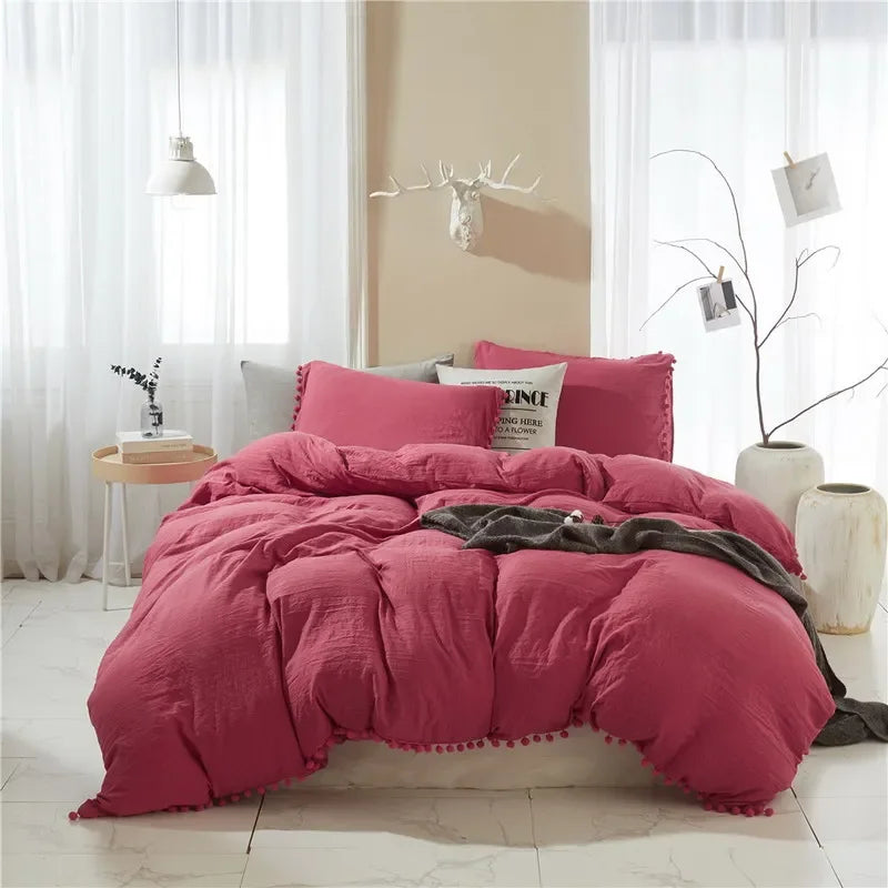 A pink duvet cover set with a pom-pom trim displayed on a bed in a bright room.