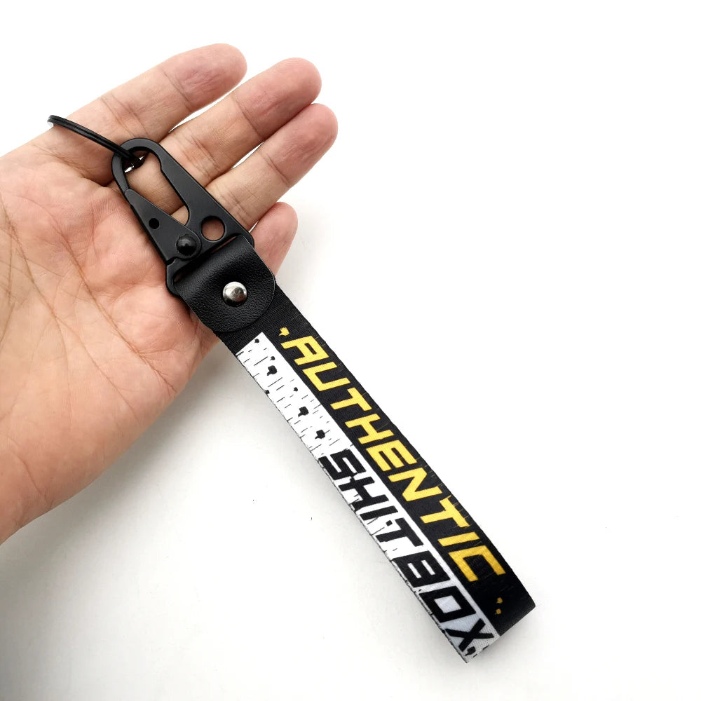 A black leather strap keychain with a white measuring tape design and the text AUTHENTIC SHITBOX is held in a hand.