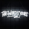A white neon sign spelling out THE WORLD IS YOURS is suspended against a dark grid background.