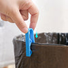 A hand is using a blue clip to secure a garbage bag onto the rim of a dark trash can.