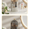 A clear glass rotating makeup brush holder with a dustproof lid displayed on a marble surface.