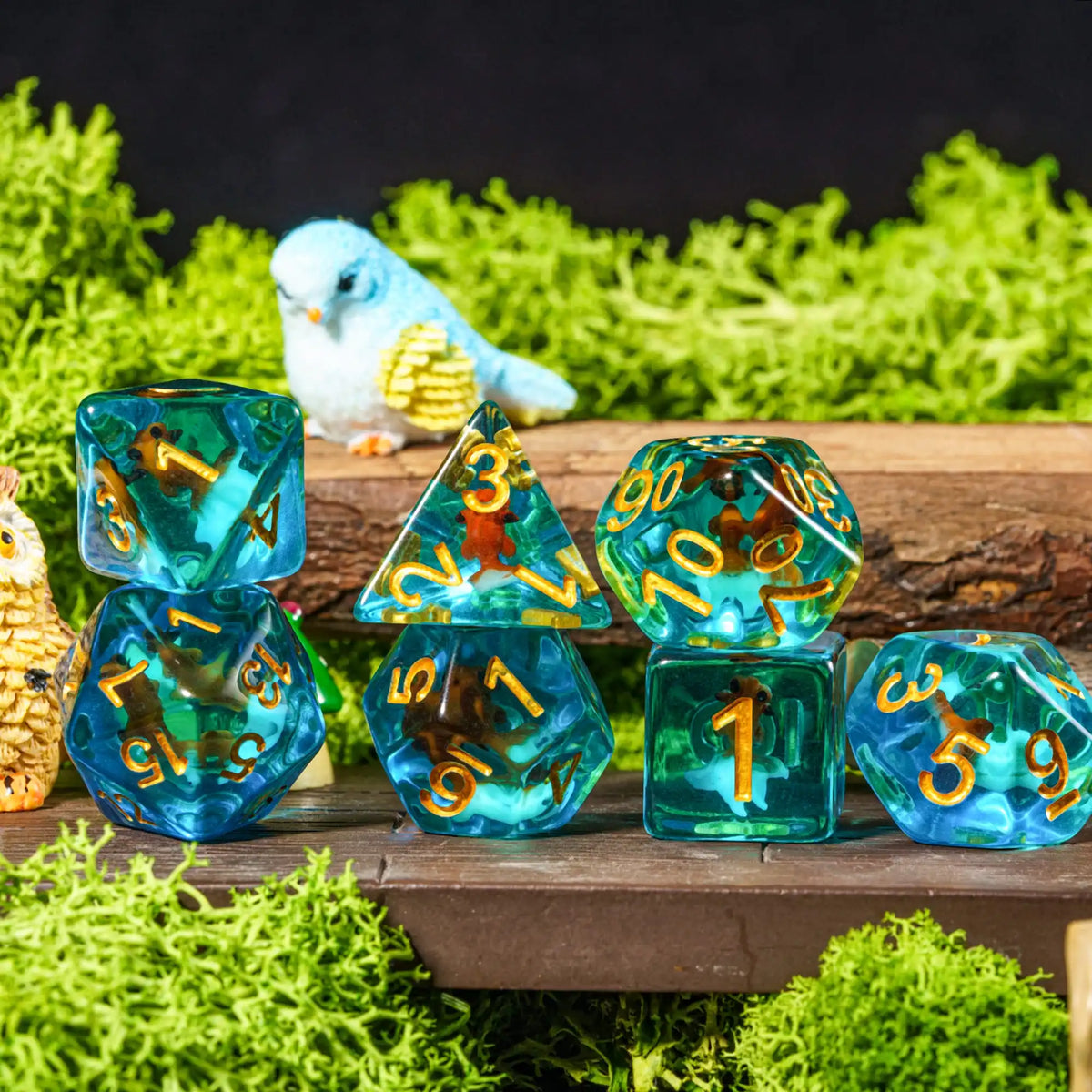 A set of colorful resin dice and a small blue bird are displayed on a wooden surface amidst green moss.
