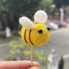 A yellow wool felt bee with white pompoms on its head is held by a finger.